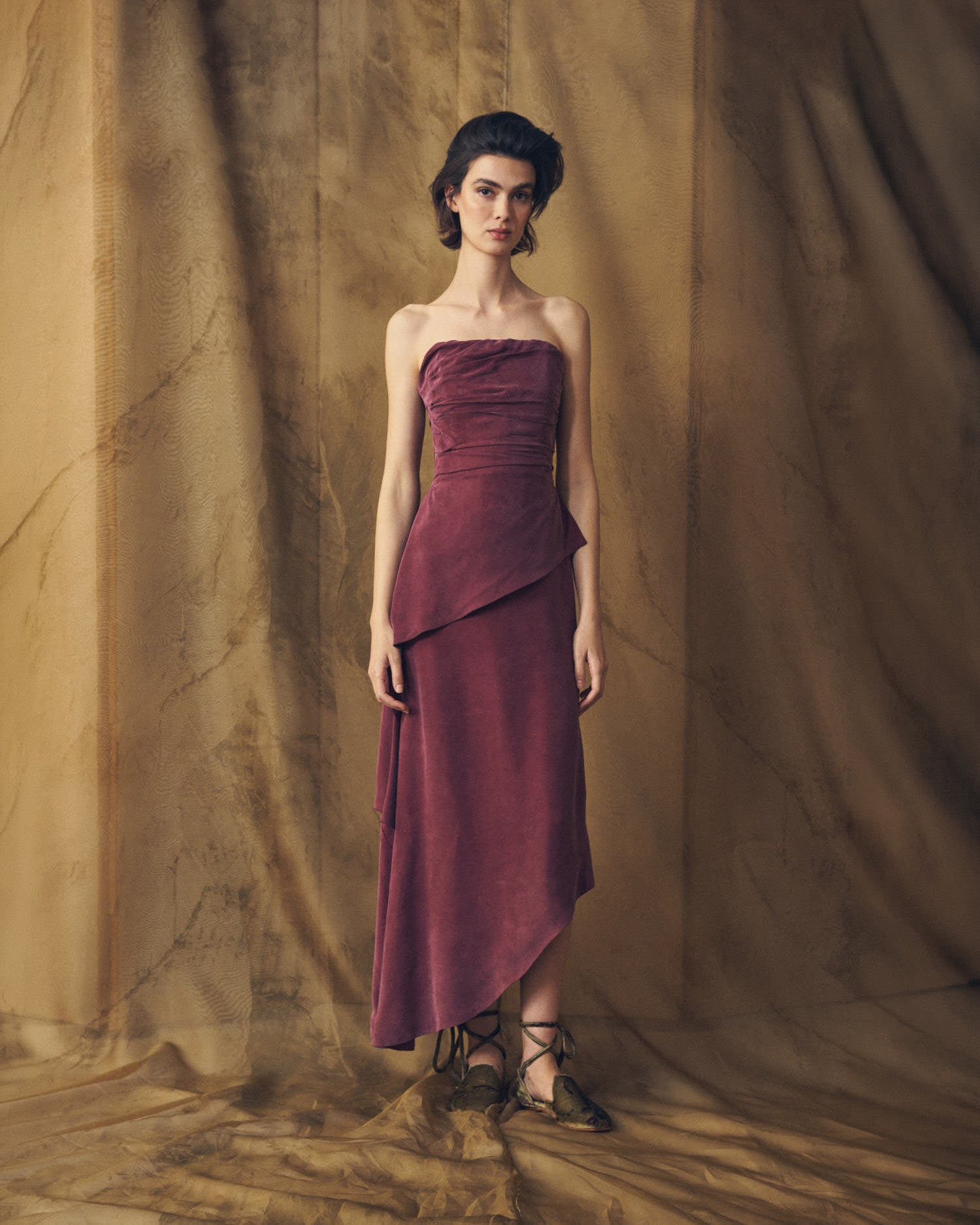 Full-length front studio view of model wearing the strapless Alina long cupro dress in burgundy with asymmetrical drape by T.ba