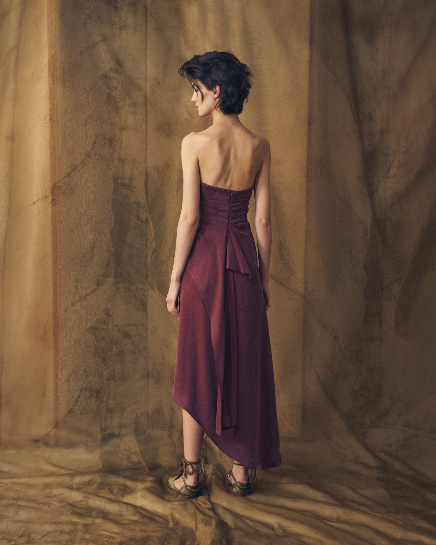 Full-length back view of model wearing the strapless Alina long cupro dress in burgundy showing back peplum drape by T.ba