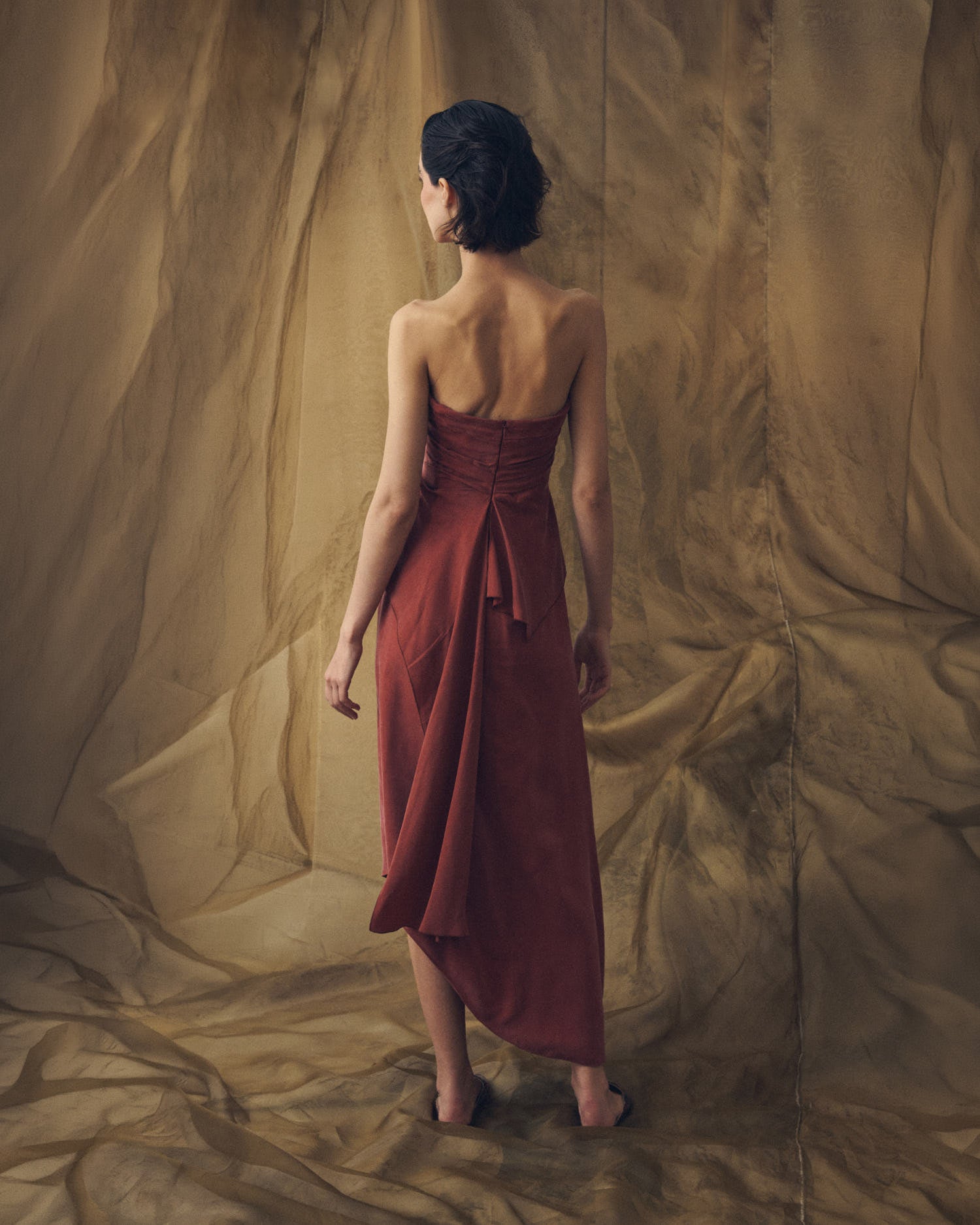Full-length back studio view of model wearing the strapless Alina maxi cupro dress in copper featuring back drape accent by T.ba