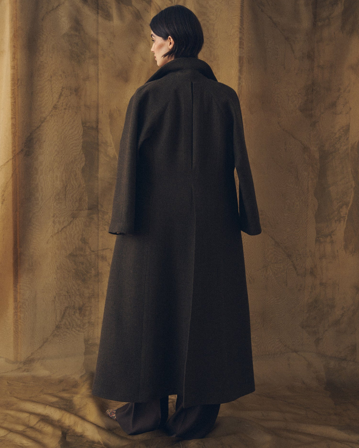 Full-body back view of the Biella Jerez long coat in brown wool-blend knit, highlighting the seamless back panel and the tailored structure of the coat, worn by a female model with dark wide-leg trousers, set against a warm textured background, T.ba.