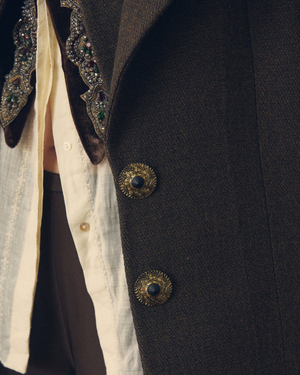 Extreme close-up view focusing on the ornate gold filigree buttons with black centers on the textured brown wool-blend fabric of the Biella Jerez long coat, also showing the textured lapel and the collar detail over a white ruffled blouse, T.ba.