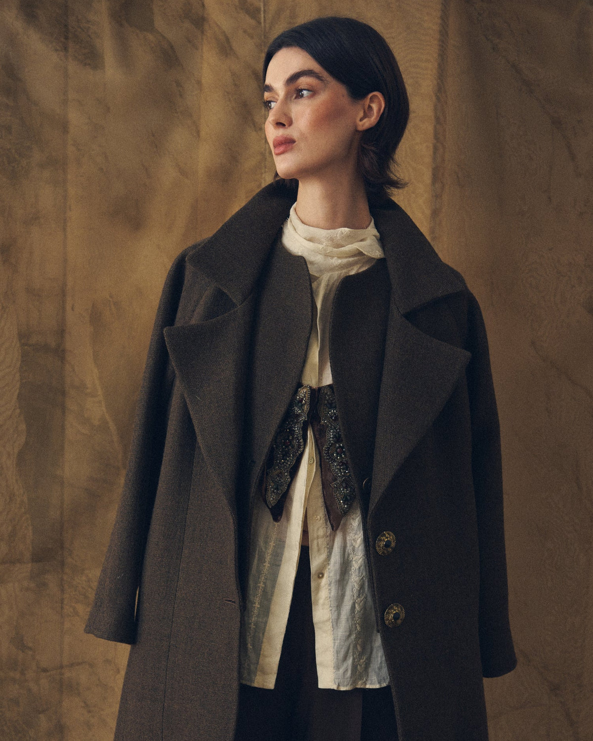 Medium front view showing the upper body and details of the Biella Jerez long coat in brown wool-blend knit, highlighting the structured collar, the open lapel, the gold filigree buttons with black centers, and the textured fabric of the coat over a delicate white ruffled blouse with a velvet bowtie, T.ba.