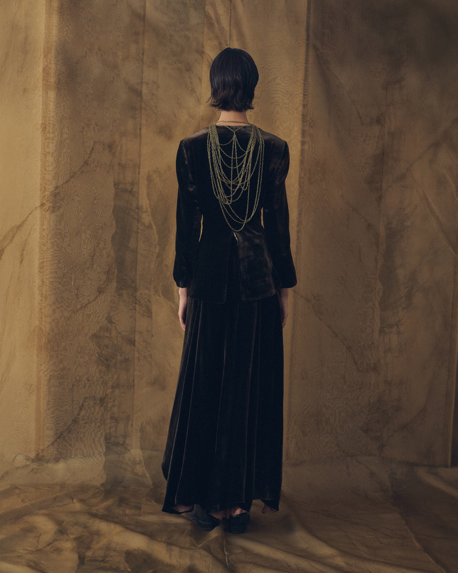 Back view of model wearing the Daria silk velvet jacket in truffle brown layered with gold chains by T.ba