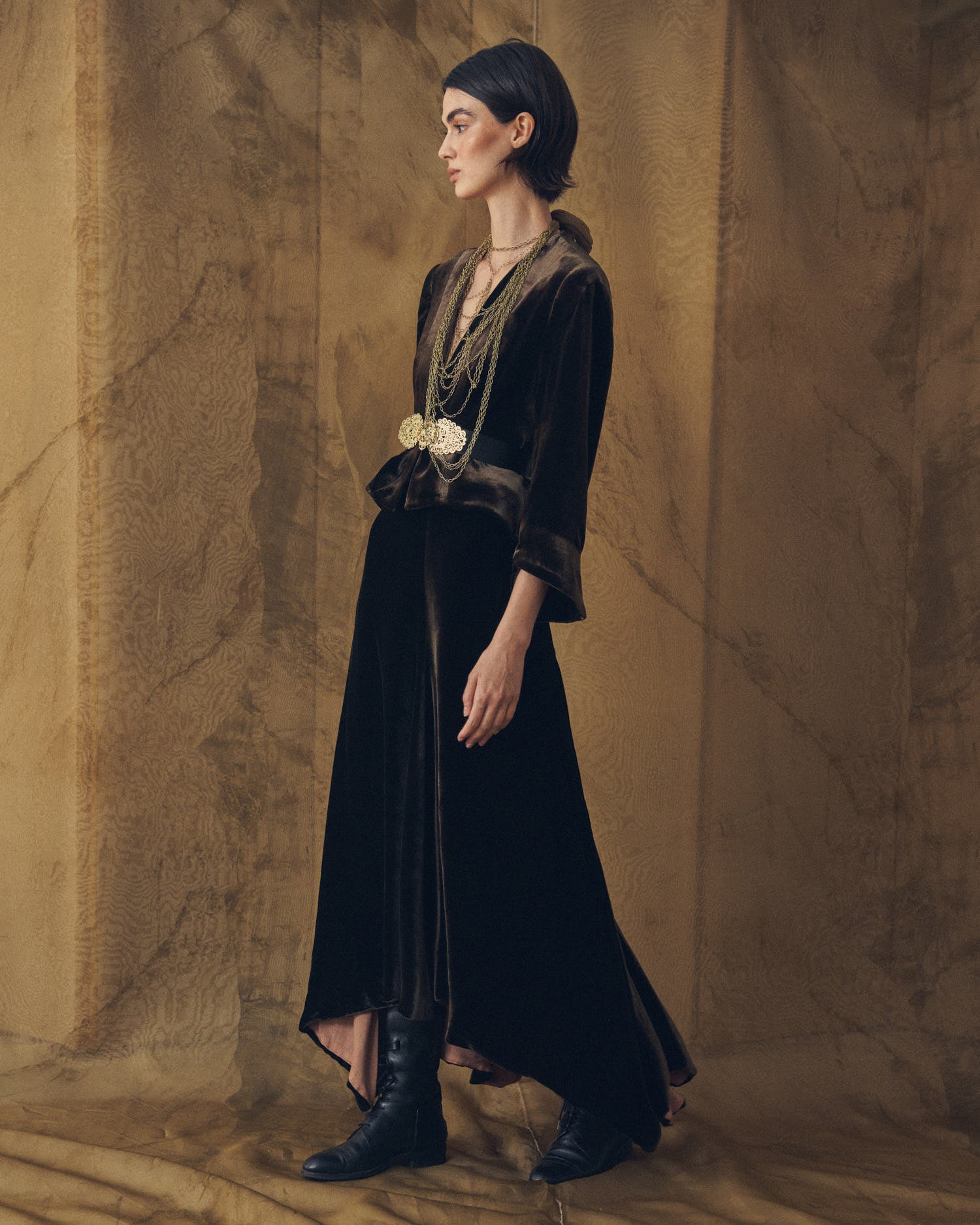 Side profile view of model wearing the Daria silk velvet jacket in truffle with long skirt and chain belt by T.ba