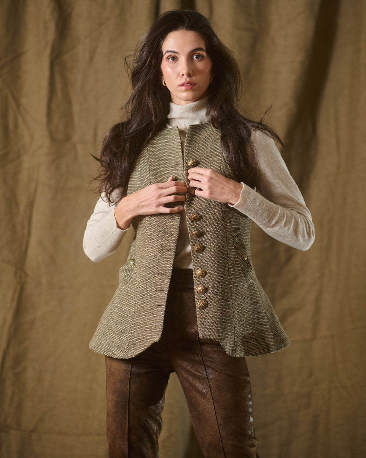 Medium close-up of model buttoning front golden buttons on taupe tweed knit Baltea vest T.ba