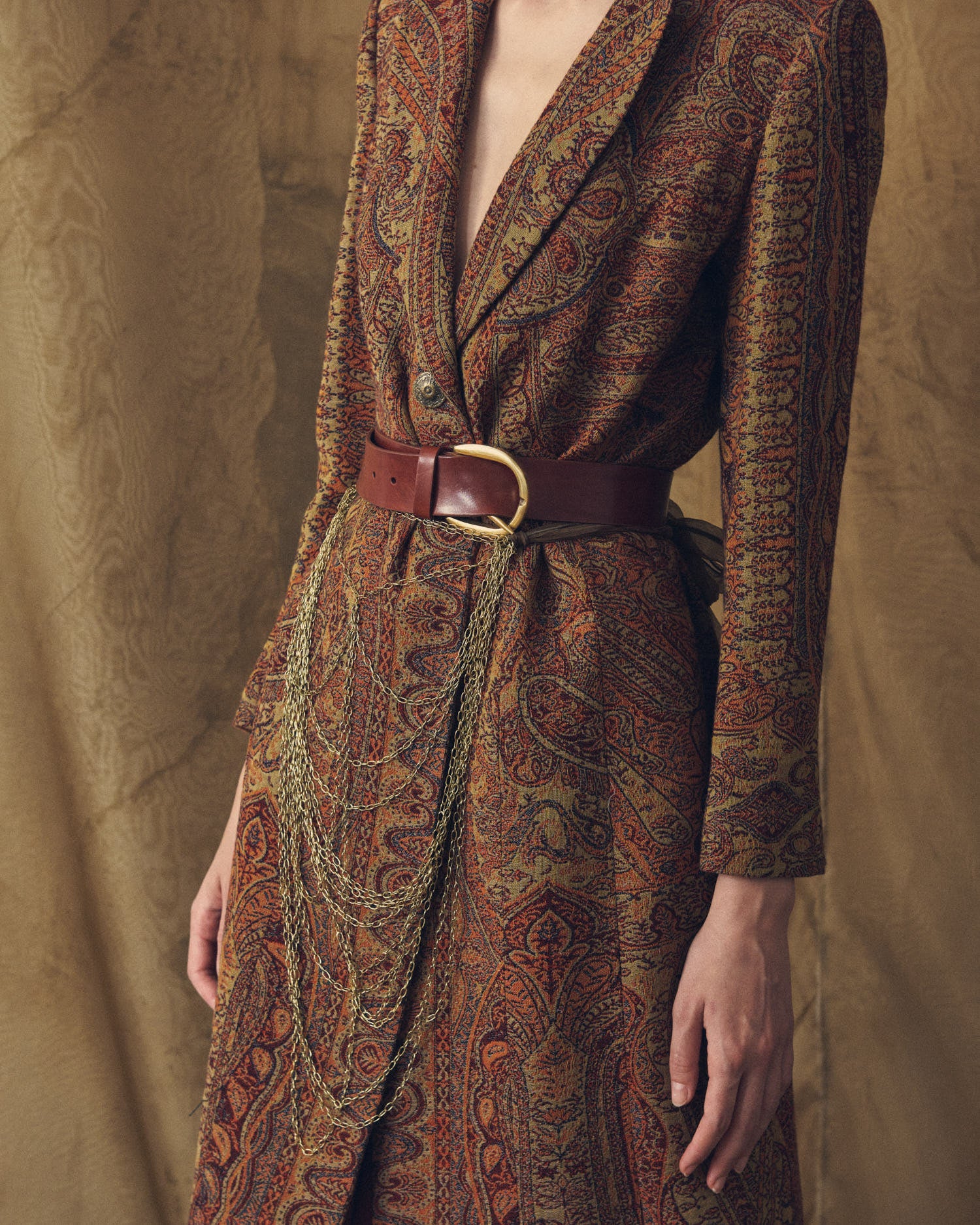 Medium shot of the Duke Belt khaki leather belt adorned with hanging gold chains over a patterned coat for women by T.ba
