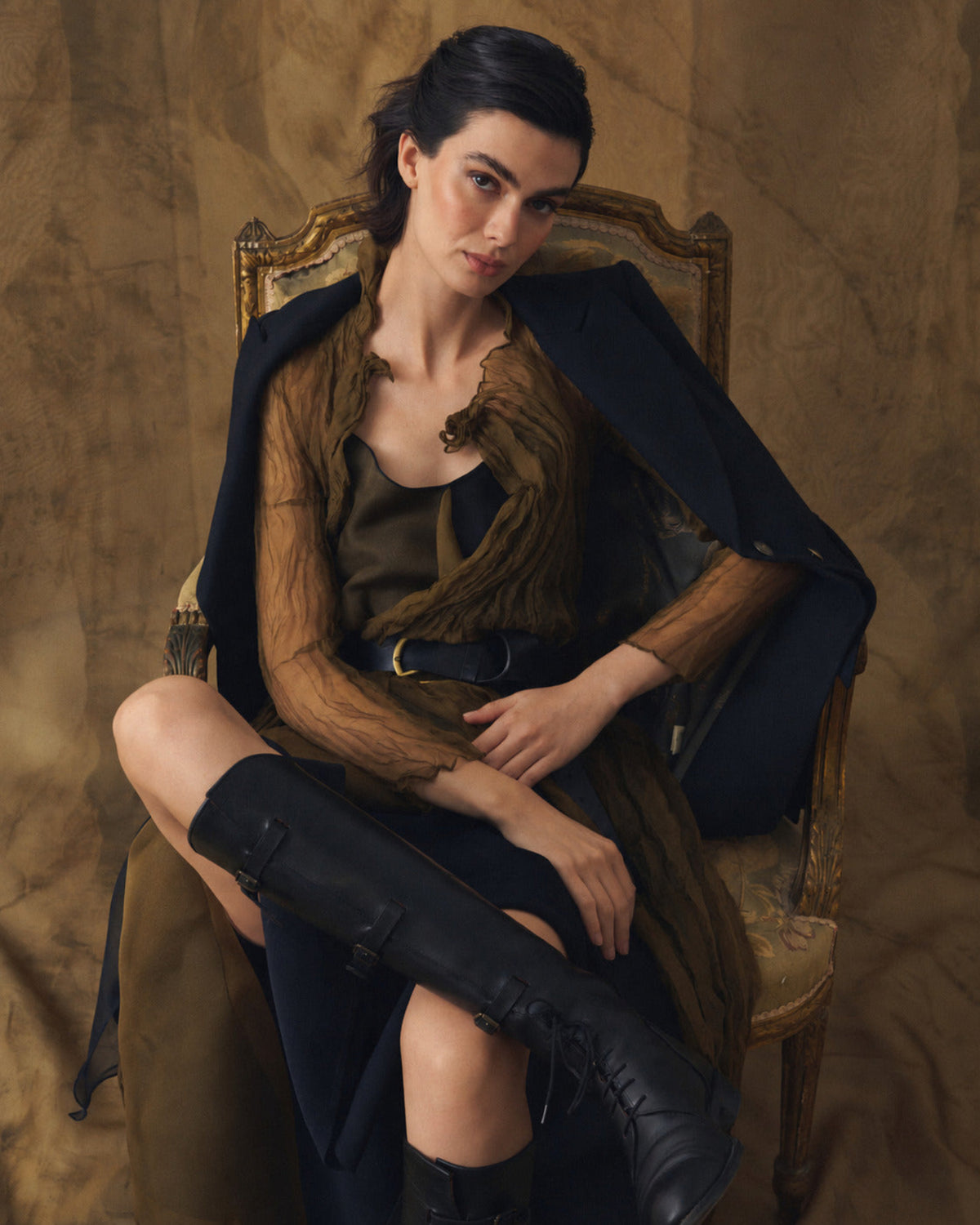 Model seated wearing black leather straps high boots paired with elegant sheer dress and jacket T.ba