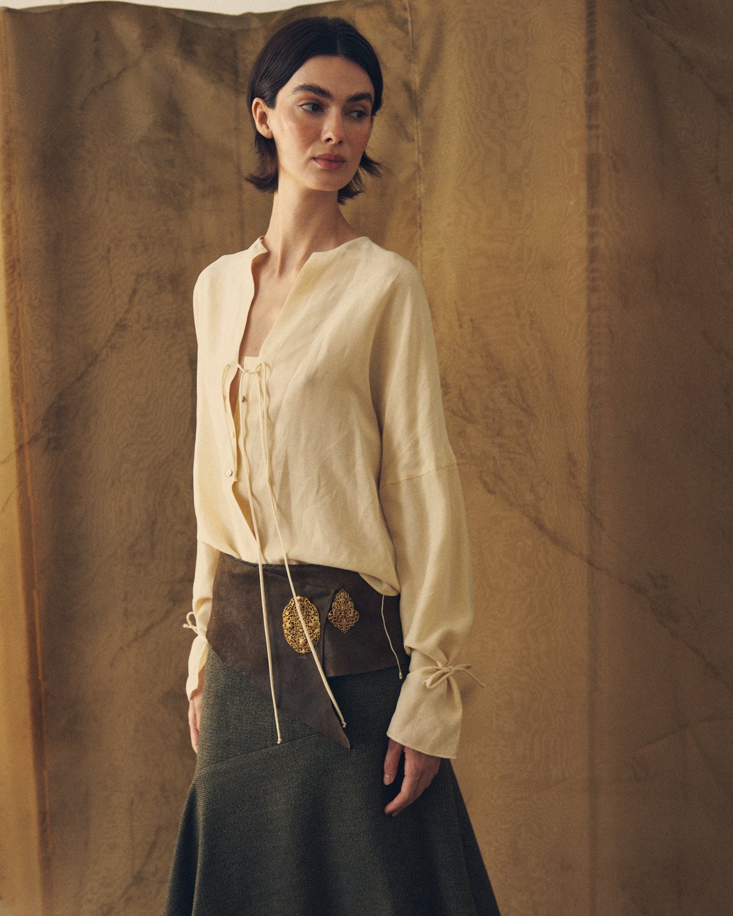 Front full-length view of the Eloise Cupro Blouse in natural cream fabric with tie details, styled with a dark skirt and leather sash belt for women T.ba