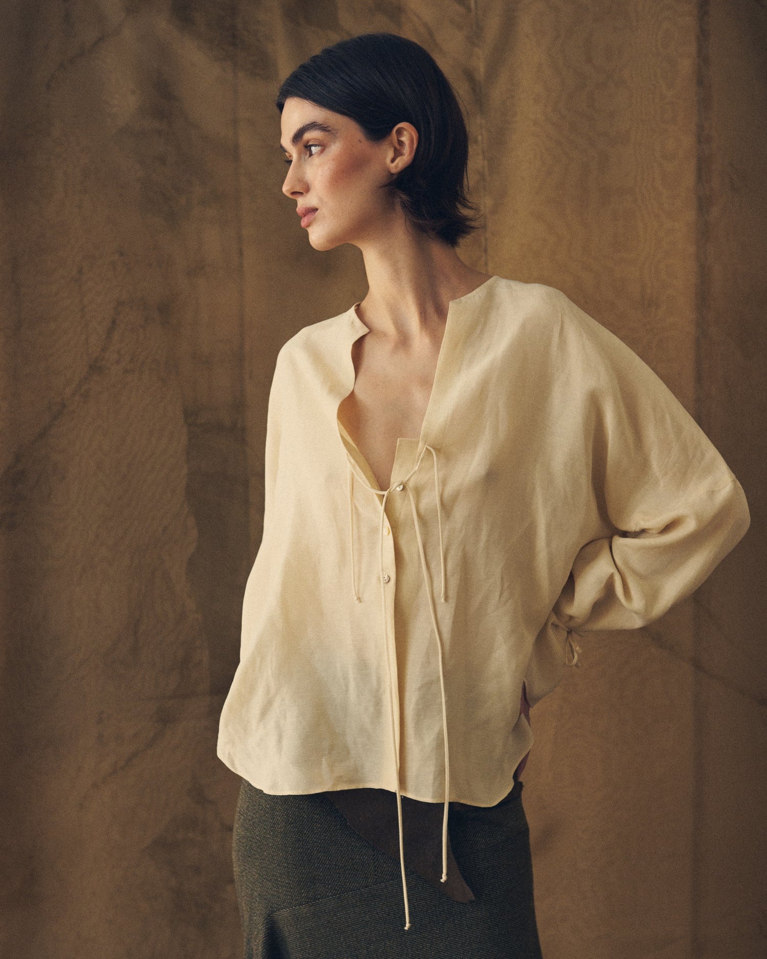 Three-quarter front view of the Eloise Cupro Blouse in natural cream featuring a split neckline with thin front ties and loose long sleeves for women T.ba