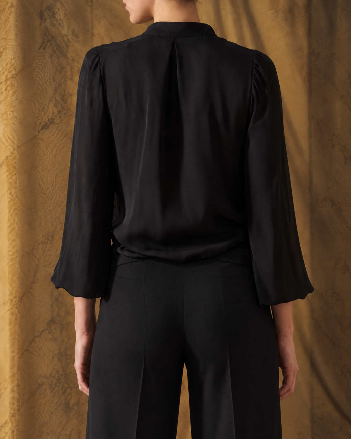 Medium back view of model wearing the Fine Crepe Toscana Blouse in black silk crepe featuring a central back pleat for women by T.ba