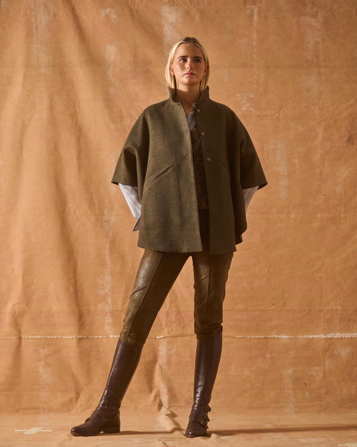 Model standing wearing chocolate leather riding boots with wool poncho coat and trousers for women T.ba