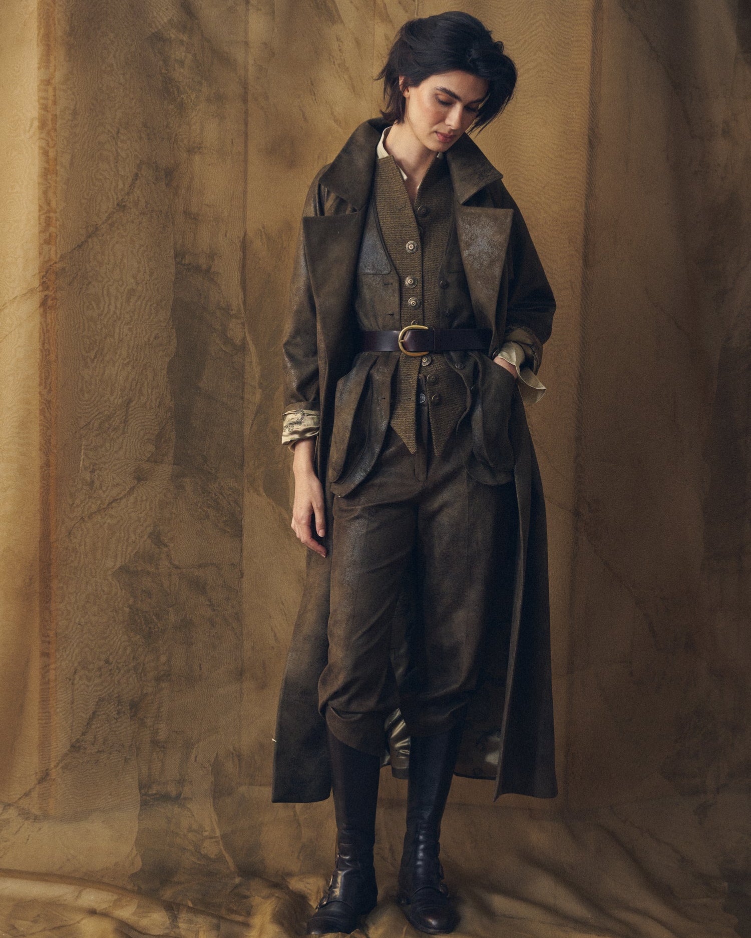 Full-length front view of a woman wearing the T.ba Gnu Biella long coat in khaki waxed linen over a vest, trousers, and tall boots.