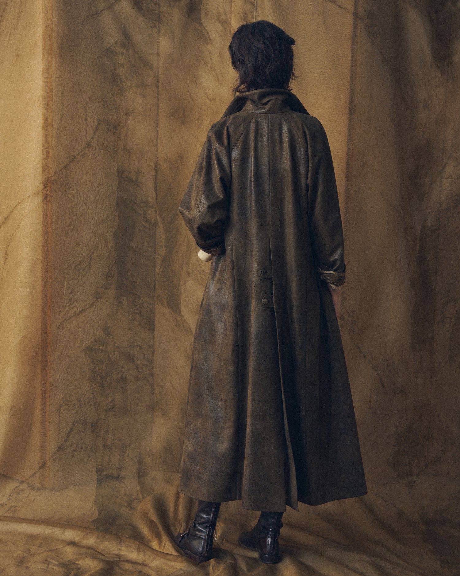 Full-length back view of a woman in the T.ba Gnu Biella long coat in khaki waxed linen, displaying its drape and back seams.