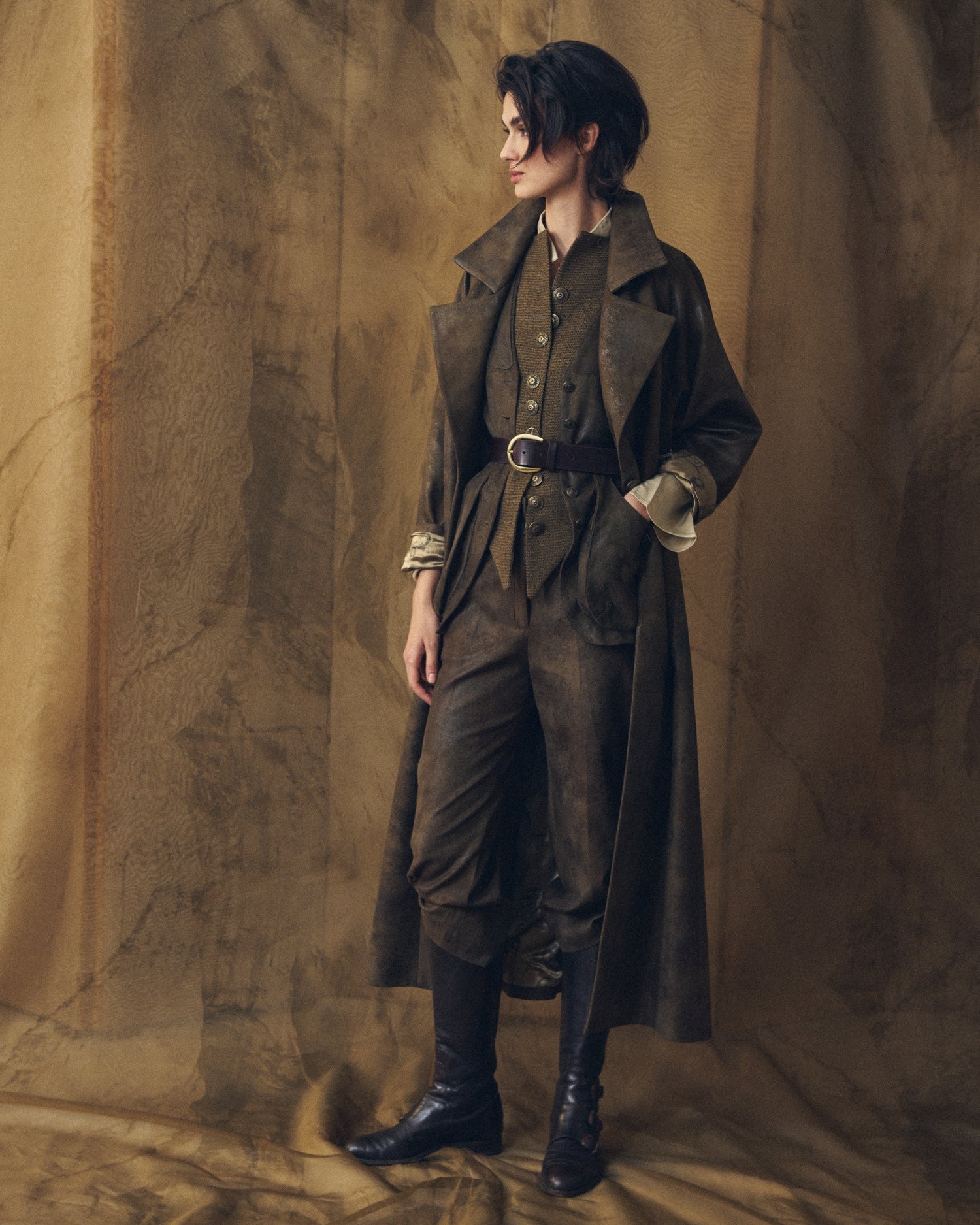 Three-quarter view of a woman wearing the Gnu Biella long coat in khaki waxed linen and full equestrian-style outfit by T.ba.