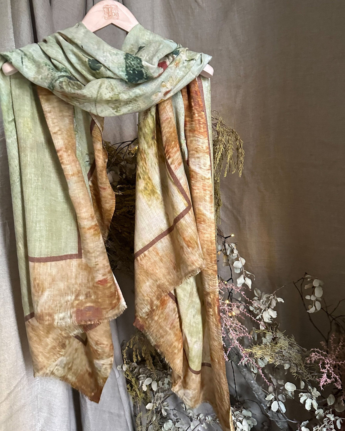 Studio shot of Greta wool shawl featuring printed green and ochre botanical pattern hanging on a wooden hanger for women T.ba
