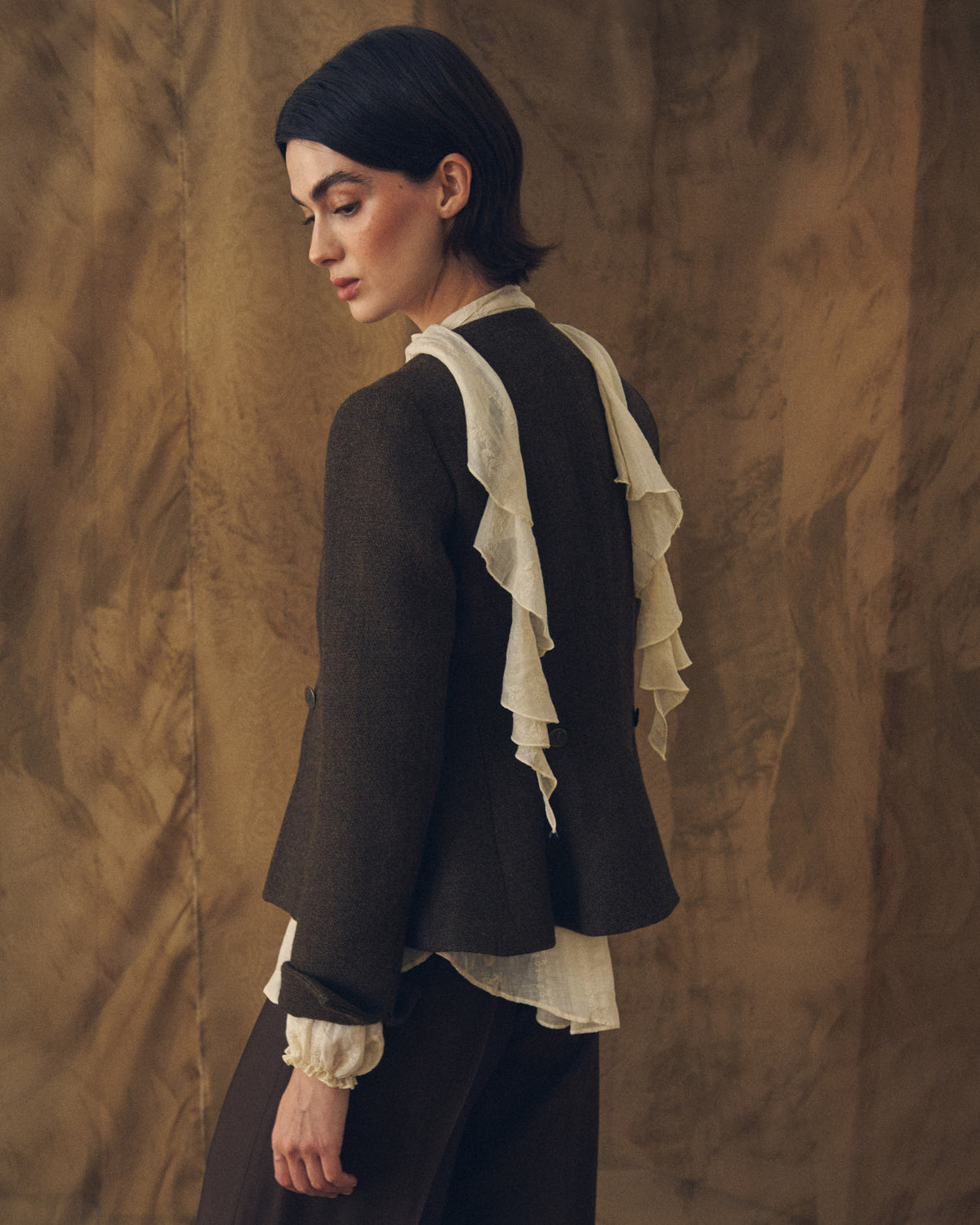 Three-quarter back view of model wearing the Jerez Greta dark brown wool jacket layered over a ruffled blouse by T.ba