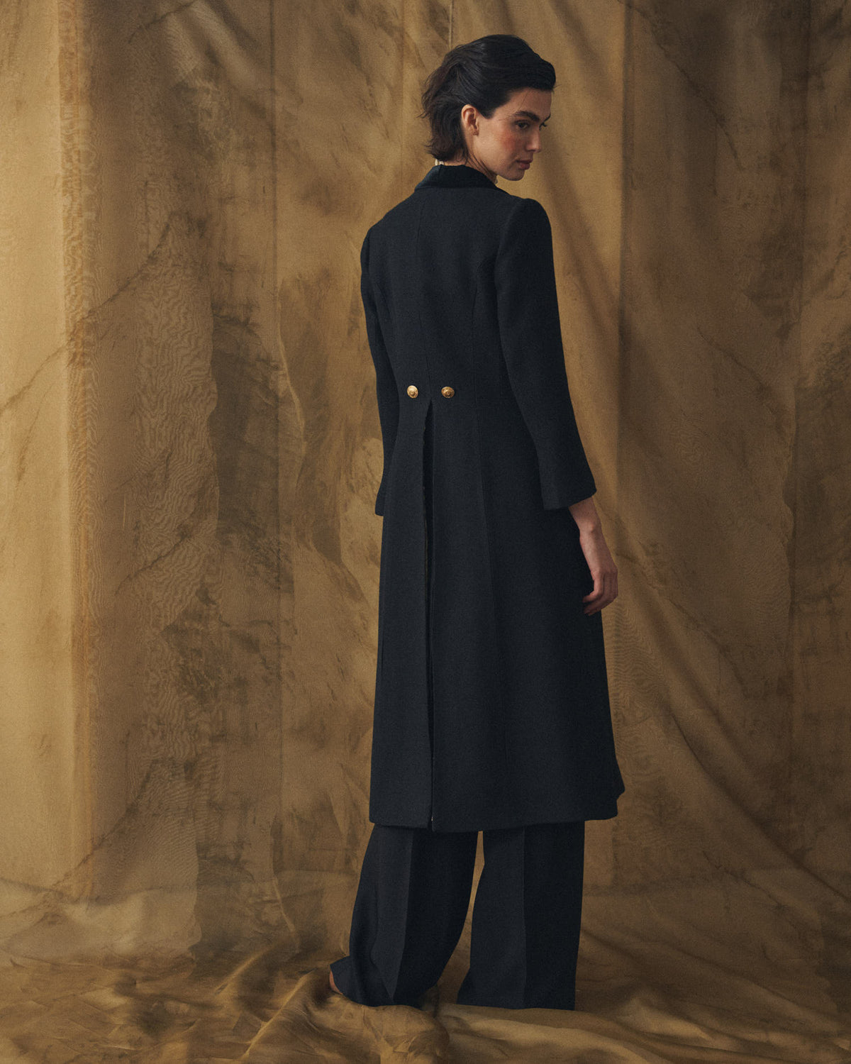 Full length back view of model wearing the Jerez Lancelot Combi Coat long black wool coat with back pleat and gold buttons for women by T.ba