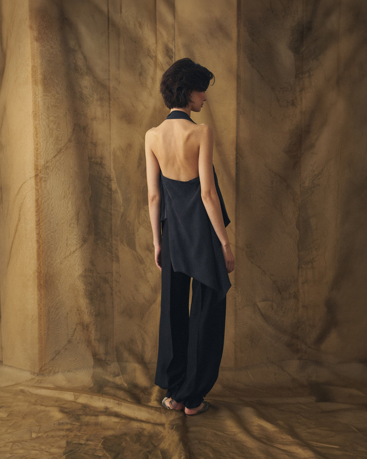 Full-length back view of model wearing the backless black Sam cupro top with draped hem by T.ba