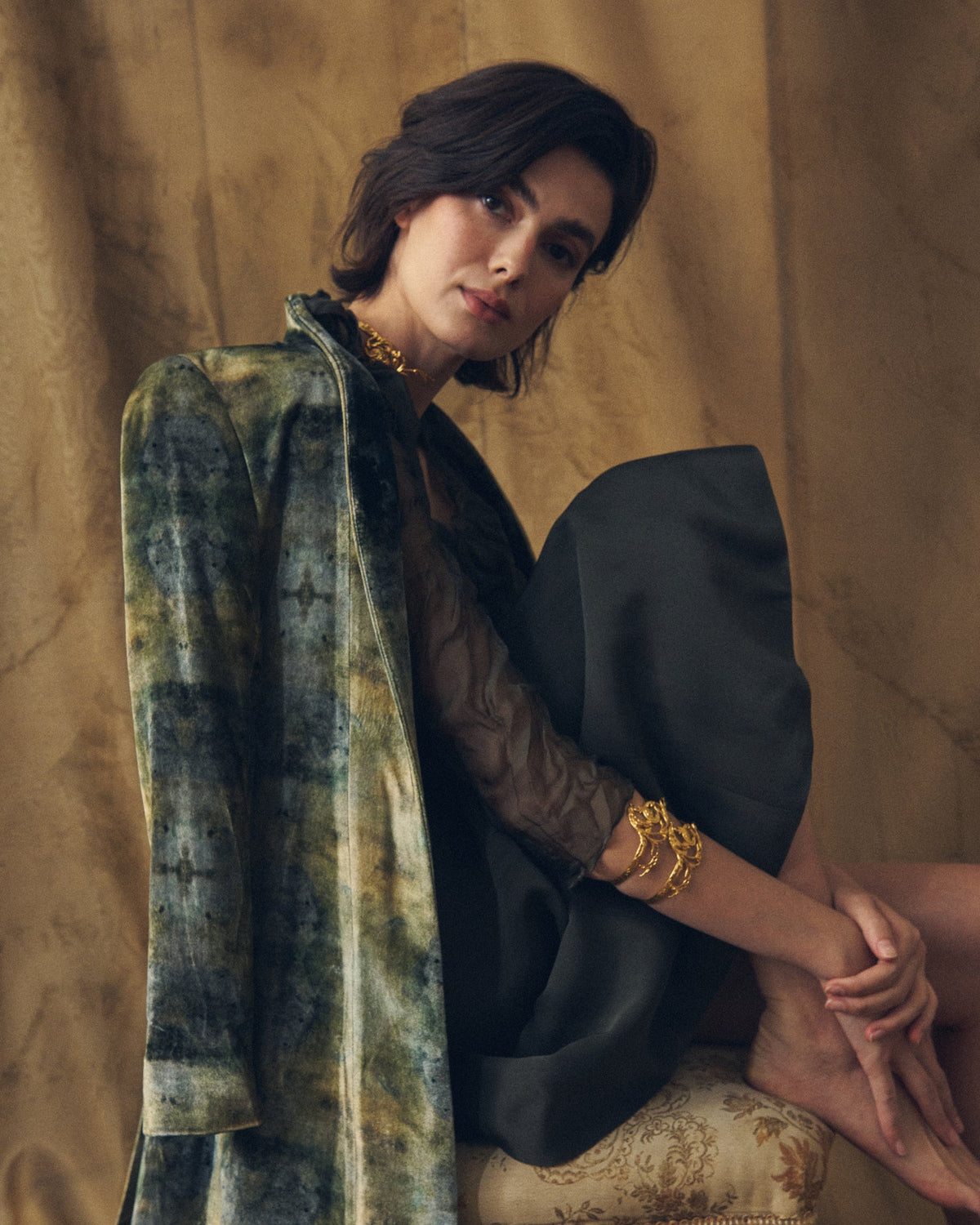 Seated model wearing Noor gold filigree cuff bracelet paired with a dark silk dress and printed coat for women T.ba