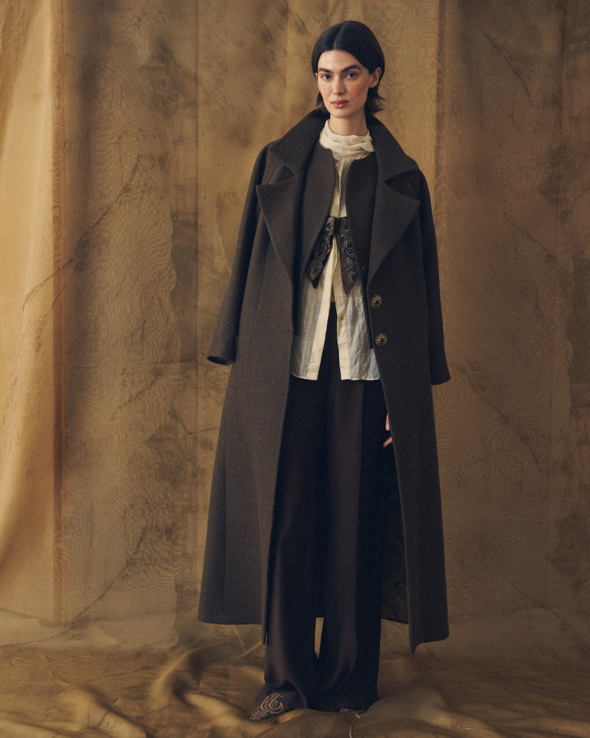 Full length view of model wearing the wide-leg Sharkskin Maxi Pants in Truffle brown layered under a long wool coat for women by T.ba
