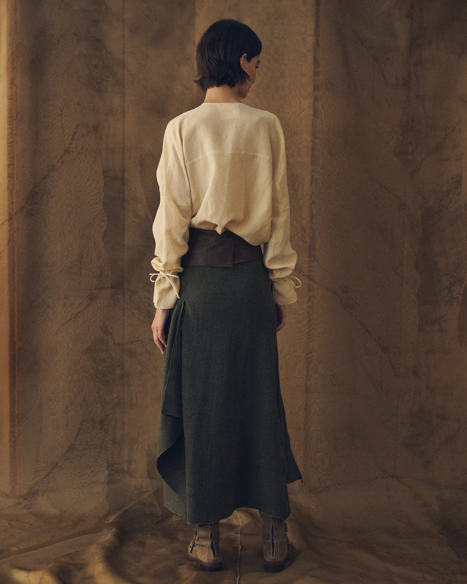 Full-length back studio view of model wearing the Toscana dark green maxi tweed skirt with asymmetrical drape by T.ba