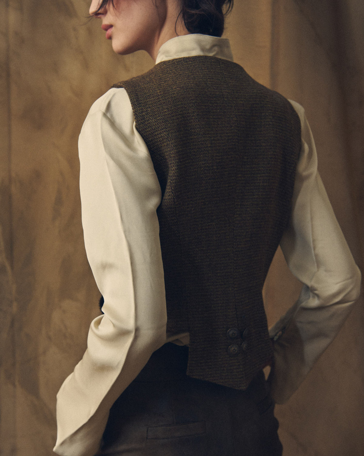 Medium rear three-quarter view showing the tailored back construction and button details of the Tweed Short Baltea Vest in brown wool for women by T.ba