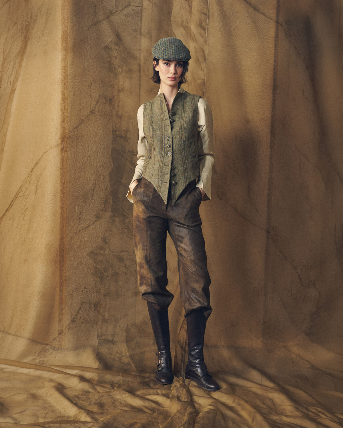 Full-length view of a model wearing the Tweed Short Baltea Vest with brown trousers, riding boots, and a cap for women by T.ba.